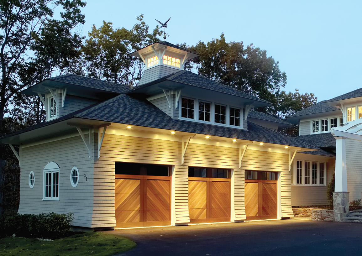 Three wood garage doors on a single-family home, at dusk, with lights on above garage doors.
